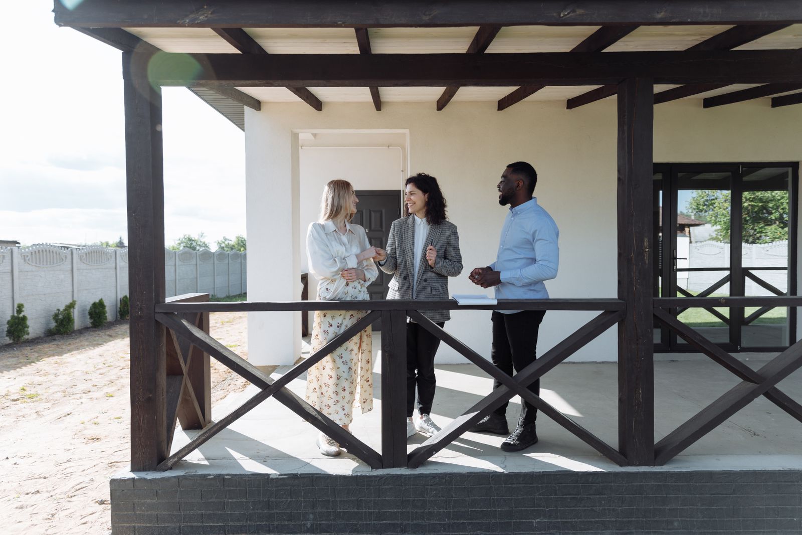 Three people in business casual clothing talking together on a covered porch of a residential house.